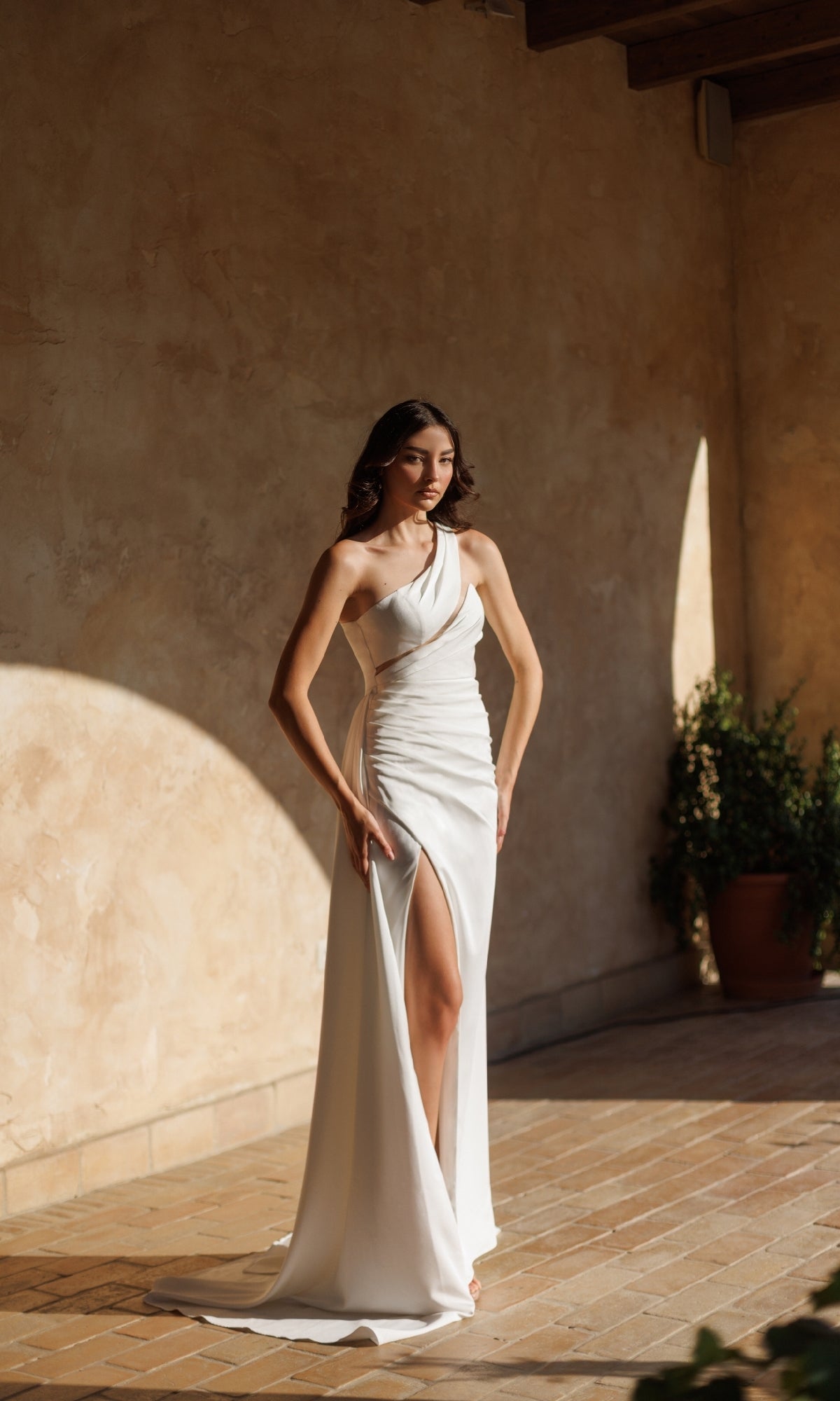 A woman stands indoors on a sunlit tile floor near a potted plant, wearing the Tina Holly TT001W—an elegant one-shoulder white prom gown with a floor-length skirt and thigh-high slit.